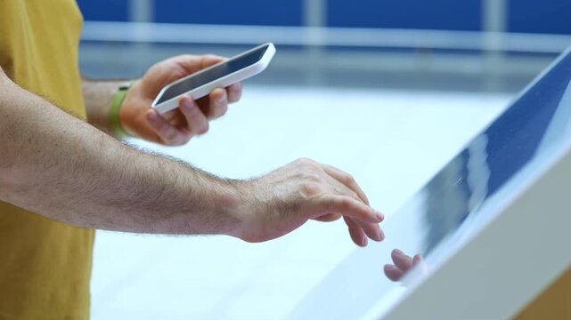 Man holds phone while touching interactive screen. Hands interact with modern display surface. Background shows indoor space with blue walls. Man wears yellow shirt and green wristband