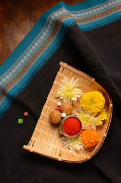 A festive Makar Sankranti photo featuring a bamboo basket holding marigold, til-gul laddus, halwa and kumkum, placed on a black traditional saree.