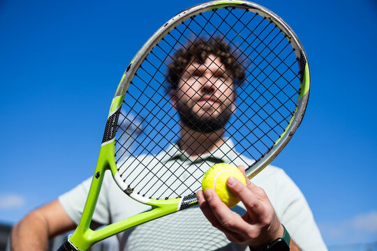 Male tennis player holding a yellow tennis ball and racket, with his face framed by the strings against a clear blue sky