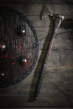 A round Viking style wooden shield with metal bosses, a black riveted rim and red paint splashes hangs beside a decorative warrior axe on a dark plank wall.