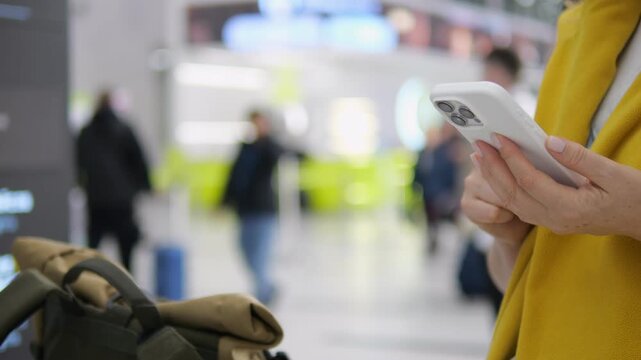 Person in yellow jacket holds phone. Background shows blurred travelers in airport. Luggage rests near foreground. People walk through terminal area. Scene captures modern travel
