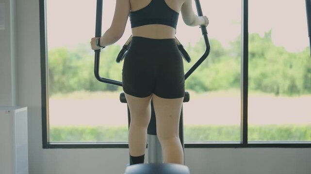 Woman exercising on elliptical machine in gym with large window and green outdoor view in bright natural light