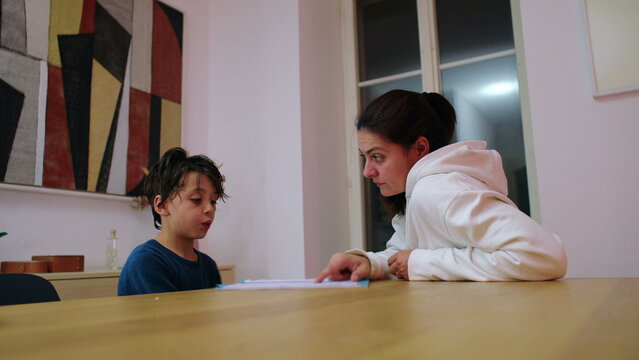 Mother supervises her child reciting school material at the home table in the evening while pointing to the worksheet, encouraging focus, comprehension and learning