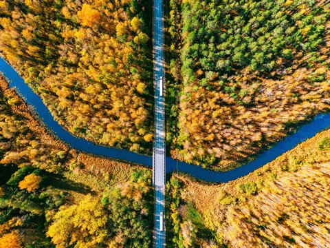 Top aerial view of commercial logistics trucks crossing bridge over winding autumn forest river
