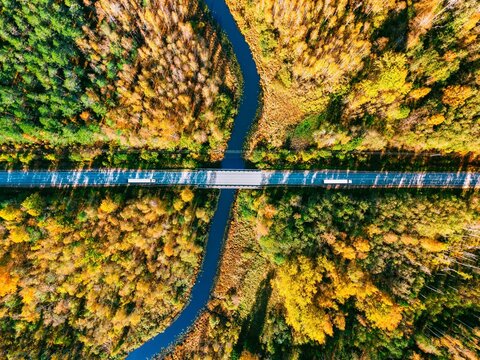 Top down aerial view of river bridge with cargo trucks on winding road in autumn forest