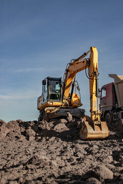 A tracked excavator rests on a mound of soil beside a dump truck at a construction site during the day