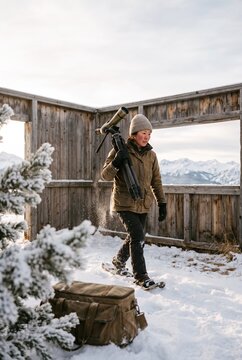 Asian woman wildlife researcher walking in snowshoes carrying a spotting scope on a tripod inside a wooden viewing blind with snowy mountains behind
