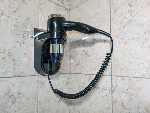 Wall mounted hair dryer with coiled cord on tiled bathroom wall. Hotel bathroom amenity. Personal grooming and hair care. Guest accommodation convenience