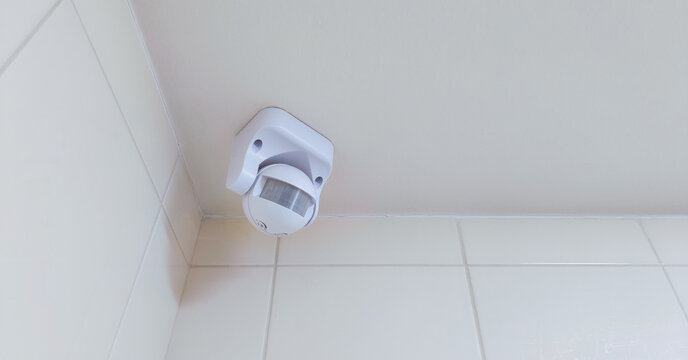 Motion sensor mounted high on tiled wall in interior corner. Automatic occupancy detection for lighting and security systems. Home automation and energy efficiency