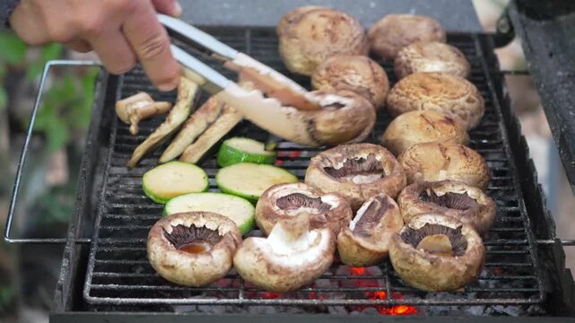 Outdoor cooking with portobello mushrooms and sliced squash on a charcoal grill during a backyard gathering. High-angle static shot.