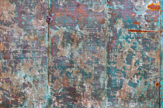 Full-frame close-up reveals a weathered metal surface featuring peeling paint in shades of brown, teal, blue, and off-white, along with significant rust. The texture showcases age and exposure to