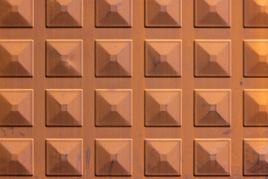 Close-up view of a textured surface featuring a grid of raised squares, each with a pyramidal indentation. The rusty brown color and visible scratches enhance the geometric relief and surface detail.