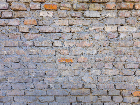Full-frame close-up of a weathered brick wall featuring rectangular bricks in gray, reddish-brown, and orange hues. Mortar fills gaps, showcasing rough texture and aged appearance.