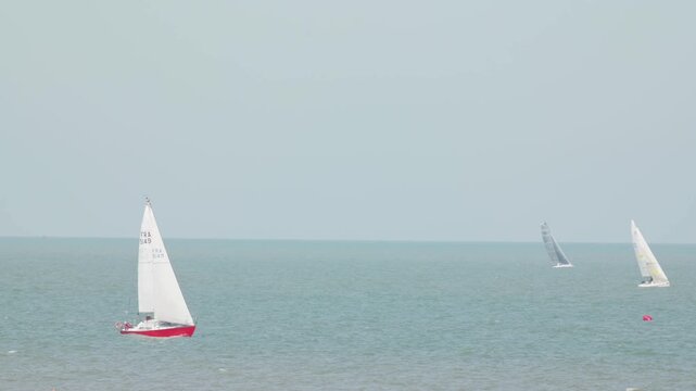 Red hulled sailboat with white sails and distant boats at sea compressed perspective long zoom shot