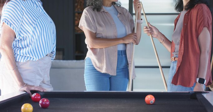 Three women holding cues and leaning on black pool table in game room, with pool balls