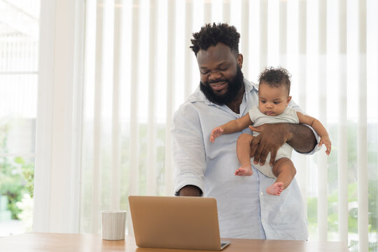 Happy African American father holding cute infant baby while working remotely on modern laptop computer at home, embracing parenting, career life, daily child care
