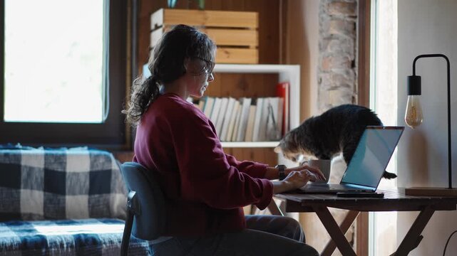 Young woman working from home in a cozy home office with her cute cat standing on the desk beside her laptop, capturing remote work and everyday life with pets.