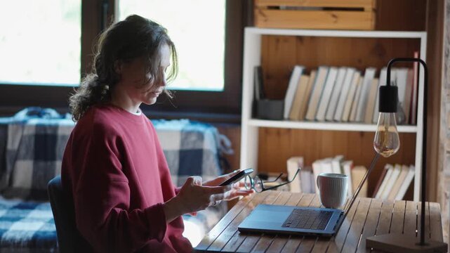 Young woman using her smartphone while sitting in front of a laptop in her cozy home office, multitasking between mobile communication and remote work.