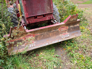 Close-up of a rusty front dozer blade on an old LKT forestry skidder, showing hydraulic cylinders, worn steel and vegetation. Heavy logging machinery used in forest operations. © Marek