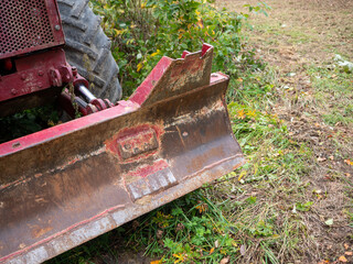 Close-up of a rusty, worn front dozer blade on an old red LKT forestry skidder. Scratched steel, hydraulic cylinder and tractor tire show heavy logging equipment in a grassy rural setting. © Marek