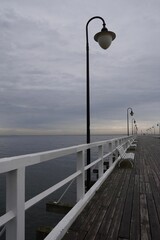 A scenic view of a wooden pier stretching into the sea on a cloudy day, featuring white railings and vintage lanterns. Perfect for travel, tourism, and melancholic nature themes. © Alina