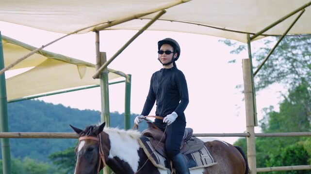 Slow Motion. Young Asian woman riding horse in outdoor arena wearing helmet and sunglasses