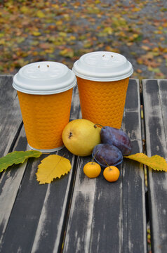 Two orange paper cups with white lids and fruits arranged on wooden table surface. Cups with hot baverage for two persons and fruits dessert in autumn park cafe .
