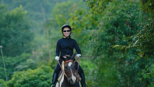 Woman wearing black helmet and riding black white horse in green outdoor field with dense tree background