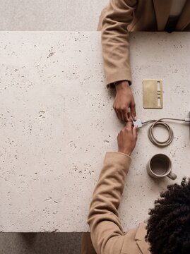 Overhead view of two colleagues in beige coats sharing a braided charging cable at a minimalist travertine stone desk with copy space