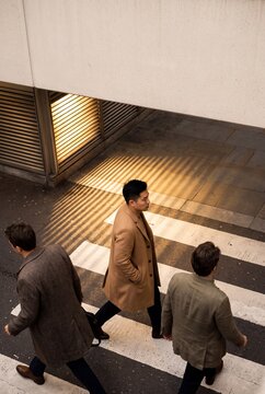 Three stylish businessmen in coats walking in opposite directions across a pedestrian crosswalk in the city during golden hour
