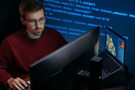 Software engineer in glasses sits in a dark room at a desk with multiple computers. Male developer works on complex data science project, programming script lines on a wall behind. Copy space