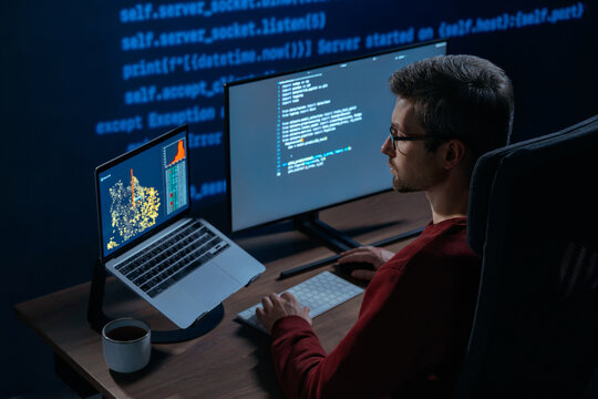 Concentrated male coder works at multi-monitor setup in a dimly lit room with lines of code projected in the background. Software engineer tackles a challenging machine learning script late at night