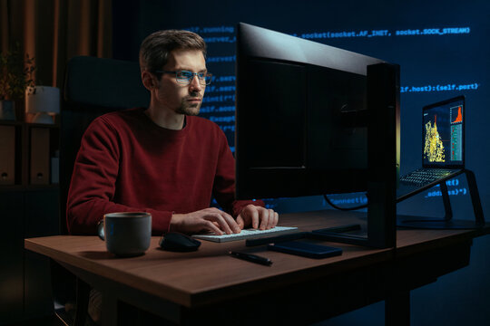 Software engineer in glasses working late, types code in a dark home office, with a coffee cup on the desk. Coder works on machine learning models, projection of programming script running behind him