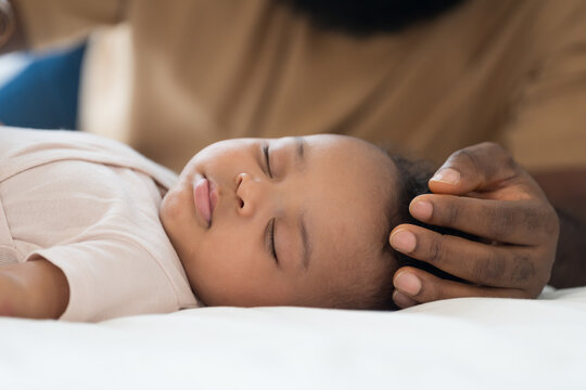 Close up of peaceful African American newborn baby sleeping soundly on white bed while loving father gently caressing child head with care, warmth and protection in bedroom at home