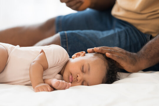 Close up of peaceful African American newborn baby sleeping soundly on white bed while loving father gently caressing child head with care, warmth and protection in bedroom at home