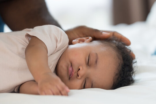 Close up of peaceful African American newborn baby sleeping soundly on white bed while loving father gently caressing child head with care, warmth and protection in bedroom at home
