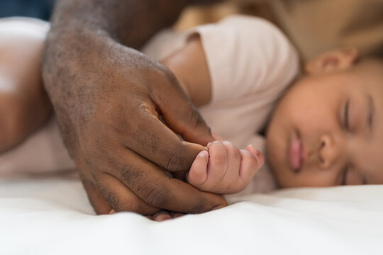 Hands of caring black father holding little hands of mixed face newborn baby infant on white bed, baby care, paternal care, fatherhood, protection and healthcare bonding at home