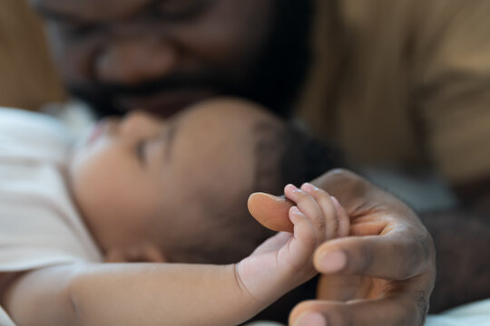Hands of caring black father holding little hands of mixed face newborn baby infant on white bed, baby care, paternal care, fatherhood, protection and healthcare bonding at home