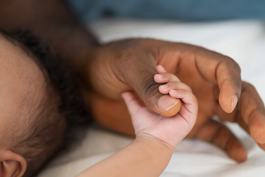 Hands of caring black father holding little hands of mixed face newborn baby infant on white bed, baby care, paternal care, fatherhood, protection and healthcare bonding at home