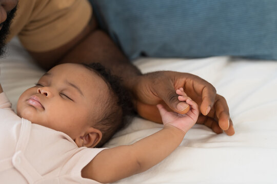 Hands of caring black father holding little hands of mixed face newborn baby infant on white bed, baby care, paternal care, fatherhood, protection and healthcare bonding at home