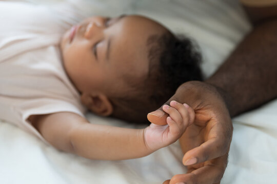 Hands of caring black father holding little hands of mixed face newborn baby infant on white bed, baby care, paternal care, fatherhood, protection and healthcare bonding at home