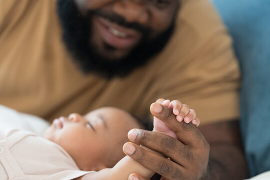 Close up of hands African American father gently holding tiny hands of newborn baby lying on white bed, baby care, paternal care, fatherhood, protection and healthcare bonding at home