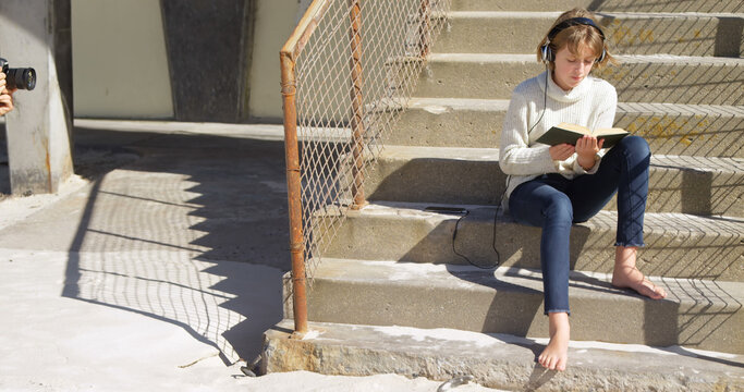 Happy caucasian girl in sweater sitting on stairs, using headphones and reading book on sunny beach