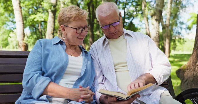 Senior couple reading book together on park bench. Focused elders enjoy quiet leisure outdoors, turning pages together. Suits retirement and healthcare messaging. Warm love theme. Peaceful.
