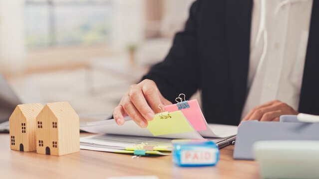 Agent reviews mortgage paperwork beside wooden house models inside office. Real estate investment financing, property tax payment assessment, home loan approval documentation.