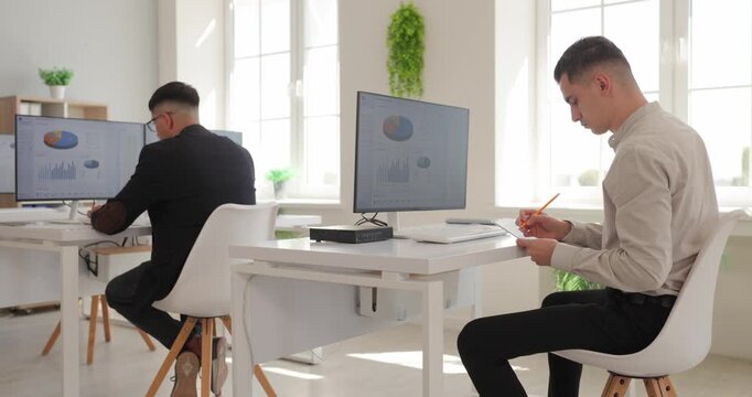 Colleagues work on computers with financial dashboards. Two businessmen review revenue charts, profit data and reports at office desks. Teamwork in corporate finance and business analytics.