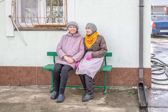 Two elderly women sitting together on a bench outside a residential building. Friendship and companionship in later life. Senior social connection and everyday community life