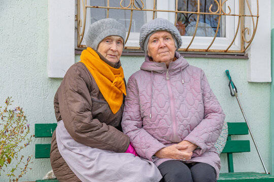 Two elderly women sitting together on a bench outside a home. Senior friendship and companionship. Aging, community and everyday life. Older adult support and wellbeing