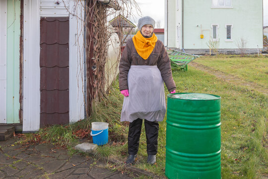 Elderly woman standing beside a green rain barrel in her yard. Senior woman tending a home garden. Gardening and outdoor maintenance. Rural living and self-sufficiency. Seasonal chores