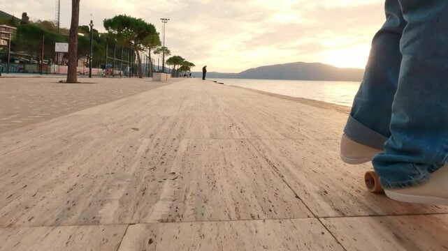Ground-level close-up shot of a skateboard rolling along a seaside promenade at sunset, showing the rider&rsquo;s feet in white sneakers, the board deck and wheels, with coastal scenery in the background.
